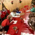 Zdjęcie przedstawia dzieci siedzące przy dużym czerwonym stole i piszą list do Świętego Mikołaja.