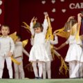 Zdjęcie przedstawia przedszkolaków występujących na scenie. Wszystkie dzieci są ubrane w jednakowe białe stroje ze złotymi skrzydełkami oraz żółtymi dodatkami.