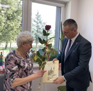 Na zdjęciu wójt, który składa życzenia jubilatce Pani Paulinie Żak z okazji 82. urodzin. Wójt wręcza Pani Paulinie czerwoną różę oraz książkę pt. "Powiat wolsztyński na dawnej pocztówce" autorstwa Artura Skorupińskiego.