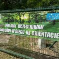 Zdjęcie przedstawia baner, na którym jest napisane: "Witamy uczestników zawodów w biegu na orientację". Na banerze jest osoba biegnąca po ścieżce wśród drzew. Logo azymut Mochy.