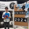 Na zdjęciu Krzysztof Kalitka pozuje z dyplomem i książką na tle tablicy korkowej, na której widnieje napis: "Dyktando 2024".