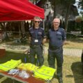 Na zdjęciu dwóch policjantów. Stoją oni przy stoisku, na którym znajdują się m.in. kamizelki odblaskowe czy odblaskowe worki na plecy. Na fotografii uchwycono kawałek wozu policyjnego.