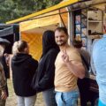 Uczestnicy wydarzenia przy foodtrucku, jeden mężczyzna uśmiecha się do aparatu.