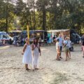 Uczestnicy wydarzenia zgromadzeni przxy foodtruckach, stoją na boisku z piasku.