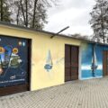 Zdjęcie przedstawia frontową część hangaru, na którym namalowano mural przedstawiający jezioro, żaglówki i dziewa