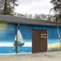 Zdjęcie przedstawia frontową część hangaru, na którym namalowano mural przedstawiający jezioro, żaglówki i dziewa