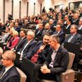Na zdjęciu widoczne są osoby siedzące w sali konferencyjnej.
