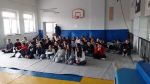 Na zdjęciu widoczni uczniowie siedzący na matach w sali gimnastycznej podczas warsztatów.