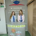 Na zdjęciu widać dwie uczennice stojące przy stanowisku ustawionym w szkole. Jest to miejsce, przy którym można odebrać dokumenty do rekrutacji.