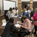 Na zdjęciu widać panią, która pomaga kobiecie przybyłej na spotkanie w wypełnieniu dokumentu. W tle znajdują się inni uczestnicy spotkania.