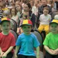Na zdjęciu trzech chłopców ubranych w kolorowe ubrania i czapki z daszkiem siedzi na widowni w sali. W tle widać inne osoby również uczestniczące w wydarzeniu.