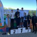 Na zdjęciu dzieci stojące na podium i obok niego z pucharami. Na fotografii obecni są także wójt gminy oraz dyrektor szkoły. Widoczne są również windery informujące o honorowym patronacie.