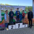 Na zdjęciu trójka dzieci stojących na podium z medalami i pucharami. Na fotografii obecni są także wójt gminy oraz dyrektor szkoły.
