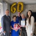 Na fotografii wójt, pracownik USC pani Natalia oraz jubilaci wraz z synem. Jubilatka trzyma bukiet kwiatów, a jubilat – list gratulacyjny. W tle widoczne złote balony z liczbą 60.