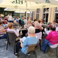 Na zdjęciu grupa seniorów spędza czas na świeżym powietrzu, siedząc przy kilku stołach ustawionych na tarasie przed restauracją. Nad nimi rozciągnięte są duże parasole chroniące przed słońcem.