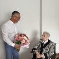 Na zdjęciu uchwycono moment, gdy wójt wręcza jubilatce bukiet kwiatów i składa życzenia.
