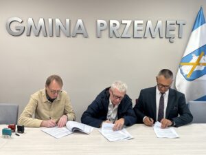 Na fotografii wójt i dwóch innych mężczyzn podczas podpisywania umowy. W tle srebrny napis: 