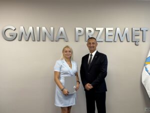 Wójt pozuje do zdjęcia z jedną z mianowanych nauczycielek. Kobieta trzyma akt mianowania w ręce. w tle srebrny napis 
