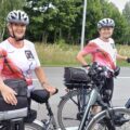 Na zdjęciu dwie uczestniczki pielgrzymki stoją obok swoich rowerów. Obie są uśmiechnięte i ubrane w koszulki z czerwonymi i białymi elementami, a na głowach mają kaski ochronne.