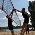 Na zdjęciu para zawodników wykonuje skok do piłki w trakcie meczu siatkówki plażowej. Jeden z graczy jest w trakcie ataku, a drugi przygotowuje się do obrony.