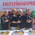 Na fotografii cztery reprezentantki KGW Starkowo pozują radośnie przed banerem akcji „Kupuj Świadomie”. Ubrane są w eleganckie czarne stroje z ludowym akcentem przy rękawach.