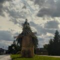 Na fotografii widać ozdobną konstrukcję przypominającą wiatrak, wykonaną z bali słomy i roślin, umieszczoną na trawie przy drodze. Konstrukcja jest bogato udekorowana roślinami, w tym ziołami, trawami i gałęziami.