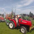 Na fotografii widać korowód dożynkowy składający się z traktorów ozdobionych roślinami. Na traktorach siedzą ludzie.
