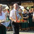 Na zdjęciu kobieta i mężczyzna, którzy niosą podarunki. Kobieta niesie chleb natomiast mężczyzna ozdobę dożynkową. Kobieta ubrana jest w strój ludowy. W tle inne osoby, które przybyły na wydarzenie.
