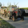 Zdjęcie przedstawia korowód dożynkowy. Na pierwszym planie traktor z przyczepą, na której siedzą ludzie.