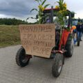 Na tym zdjęciu widzimy udekorowany traktor. Z przodu pojazdu zamocowany jest kartonowy plakat z ręcznie napisanym hasłem: „Posprzątałeś z pola chłopie, czas pomyśleć o urlopie”. Traktor ozdobiony jest łodygami kukurydzy i słonecznikami, które wystają po bokach oraz balonami. W tle widać kolejny traktor oraz ludzi, prawdopodobnie uczestników pochodu dożynkowego lub mieszkańców wsi.