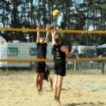 Na zdjęciu para zawodników wykonuje skok do piłki w trakcie meczu siatkówki plażowej. Jeden z graczy jest w trakcie ataku, a drugi przygotowuje się do obrony.