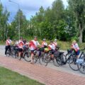 Na zdjęciu widać grupę rowerzystów, którzy pozują z rowerami na ścieżce. Uczestnicy ubrani są w jednolite koszulki z czerwonymi i białymi elementami.