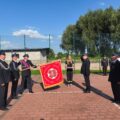 Na zdjęciu widać strażaków z Ochotniczej Straży Pożarnej (OSP) podczas ceremonii z udziałem sztandaru. Kilku strażaków stoi w szeregu, trzymając flagę z herbem OSP. Flaga jest czerwona z napisem „Bogu na chwałę, ludziom na ratunek”. W centrum zdjęcia znajduje się młoda kobieta, która trzyma kosz z akcesoriami związanymi z ceremoniałem.