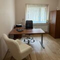 Zdjęcie przedstawia wnętrze gabinetu pedagoga z biurkiem, krzesłem obrotowym i fotelem. Pokój jest nowoczesny, z minimalistycznym wyposażeniem i naturalnym światłem, które wpada przez okno. Na biurku znajduje się laptop i drukarka.