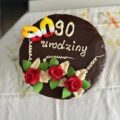 Na zdjęciu widoczny jest okrągły tort urodzinowy pokryty gładką, błyszczącą polewą czekoladową. Na jego górze, z białego kremu, widnieje napis: „90 urodziny” – litery są ręcznie napisane i lekko wypukłe. W lewym górnym rogu tortu znajdują się dwie kolorowe świeczki urodzinowe tworzące liczbę 90. Na dolnej połowie tortu znajdują się dekoracje z masy cukrowej w kształcie trzech czerwonych różyczek.
