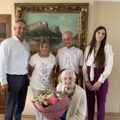 Wspólne zdjęcie, na którym znajduje się wójt, pracownik urzędu oraz jubilatka wraz ze swoimi bliskimi. Jubilatka trzyma bukiet kwiatów.