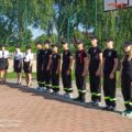 Na zdjęciu widać grupę strażaków, zarówno dorosłych, jak i młodszych członków Ochotniczej Straży Pożarnej (OSP), ustawionych w szeregu. Po lewej stronie stoją kobiety w mundurach strażackich, a po prawej grupka młodych strażaków.