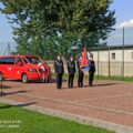 Na zdjęciu widoczna jest uroczystość strażacka z udziałem nowego pojazdu. Przed czerwonym wozem strażackim stoją czterej strażacy, trzymający sztandar z logo straży. Strażacy są ubrani w mundury, a na ich piersiach widoczna jest odznaka.