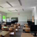 Widzimy klasę komputerową, w której znajdują się biurka z monitorami i krzesłami. W tle widać tablicę interaktywną.