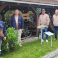 Na zdjęciu widzimy grupę czterech mężczyzn, wśród których znajduje się wójt. Mężczyźni stoją obok sztucznych ryb, które są częścią ekspozycji w Ośrodku Edukacji Przyrodniczej w Chalinie. W tle widoczna jest drewniana konstrukcja, przypominająca wiejską chatę, a wokół rośnie bujna zieleń.