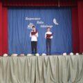 Na zdjęciu znajdują się dwaj uczniowie, stojący na scenie udekorowanej niebieską kotarą z napisem „Rozpoczęcie Roku Szkolnego”. Chłopcy ubrani są odświętnie, trzymają mikrofony i teczki z tekstem – prawdopodobnie prowadzą część oficjalną uroczystości. W tle widnieją symbole globusa, książek i pióra, podkreślające edukacyjny charakter wydarzenia.