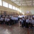 Na zdjęciu znajduje się cała społeczność szkolna, zgromadzona w sali gimnastycznej podczas apelu inauguracyjnego. Uczniowie, nauczyciele i rodzice stoją w podziale na klasy. Wszyscy ubrani są odświętnie – w białe koszule, ciemne spodnie i spódnice.
