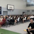 Zdjęcie przedstawia salę gimnastyczną wypełnioną uczniami i nauczycielami siedzącymi na krzesłach. Wszyscy są elegancko ubrani.