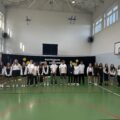 Na zdjęciu widać uczniów ustawionych w rzędzie wraz z nauczycielką na tle dekoracji z napisem: „Rozpoczęcie Roku Szkolnego 2025/2026”. Wszyscy są ubrani w eleganckie stroje. Zdjęcie zostało zrobione w sali gimnastycznej.