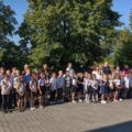 Na zdjęciu widoczna grupa dzieci, która stoi na zewnątrz w rzędzie, ubrane w odświętne stroje – głównie białe koszule i ciemne spódnice lub spodnie. Towarzyszą im nauczyciele oraz rodzice.