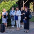 Na zdjęciu znajduje się grupa kobiet stojących na zewnątrz. Jedna z nich to dyrektor Patrycja Kmieciak, która przemawia przez mikrofon. Obok niej stoją inne kobiety, trzymające teczki i dokumenty. Przed nimi ustawiony jest głośnik. W tle widać drewnianą altanę oraz ogrodzenie.
