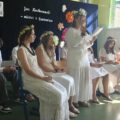 Na zdjęciu widać grupę uczniów siedzących na krzesłach w sali gimnastycznej. Są ubrani w białe stroje, niektóre osoby mają na głowach wianki z kwiatów. Jedna z uczennic stoi z mikrofonem i czyta fragmenty poezji Jana Kochanowskiego. Tło stanowi granatowa kurtyna, która jest udekorowana papierowymi kwiatami oraz widnieje na niej napis "Jan Kochanowski - mistrz z Czarnolasu".