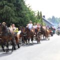 To zdjęcie przedstawia korowód dożynkowy. Grupa ludzi bierze udział w tradycyjnej paradzie, w której kluczową rolę odgrywają konie zaprzęgnięte do wozów. W tle widać osoby na motocyklach, które również uczestniczą w korowodzie.