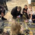 Na zdjęciu dzieci w wieku przedszkolnym siedzą wokół makiety skrzyżowania, po której poruszają się małe roboty imitujące pojazdy. Na „drodze” umieszczono znaki zakazu, przejścia dla pieszych i sygnalizację świetlną – wszystko po to, by najmłodsi mogli uczyć się zasad ruchu drogowego w praktyce. Nad przebiegiem zajęć czuwają starsi uczniowie, którzy tłumaczą zasady i pomagają dzieciom w obsłudze robotów. Na fotografii widoczny także wójt gminy, który obserwuje zajęcia.
