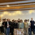 Wspólne zdjęcie uczniów zaangażowanych w projekt wraz z dyrektorem szkoły. Dwójka uczennic trzyma korkową tablicę, na której widoczne są grafiki związane z tematyką projektu.